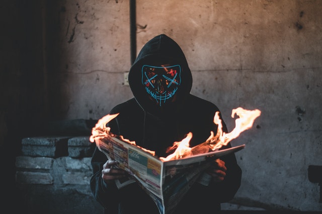 homem encapuzado lê um jornal que está pegando fogo, simbolizando os perigos das fake news e desinformação